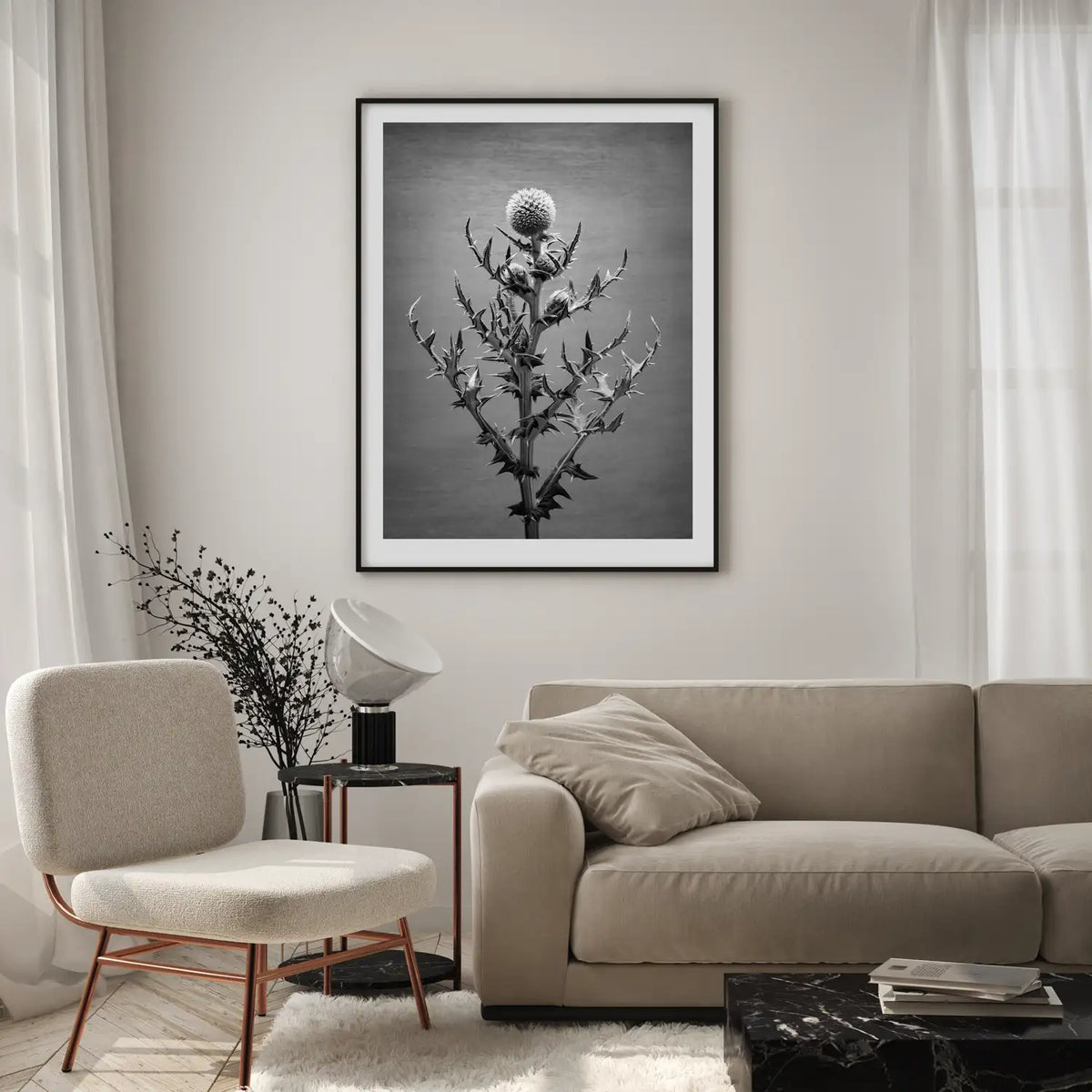 A lifestyle image for representation only showing a neutral environment of a beige lounge with chair, side table and spotlight lamp, black dried flowers with a black marble coffee table featuring a black and white photographic print with black frame of an echninops 'arctic glow'.