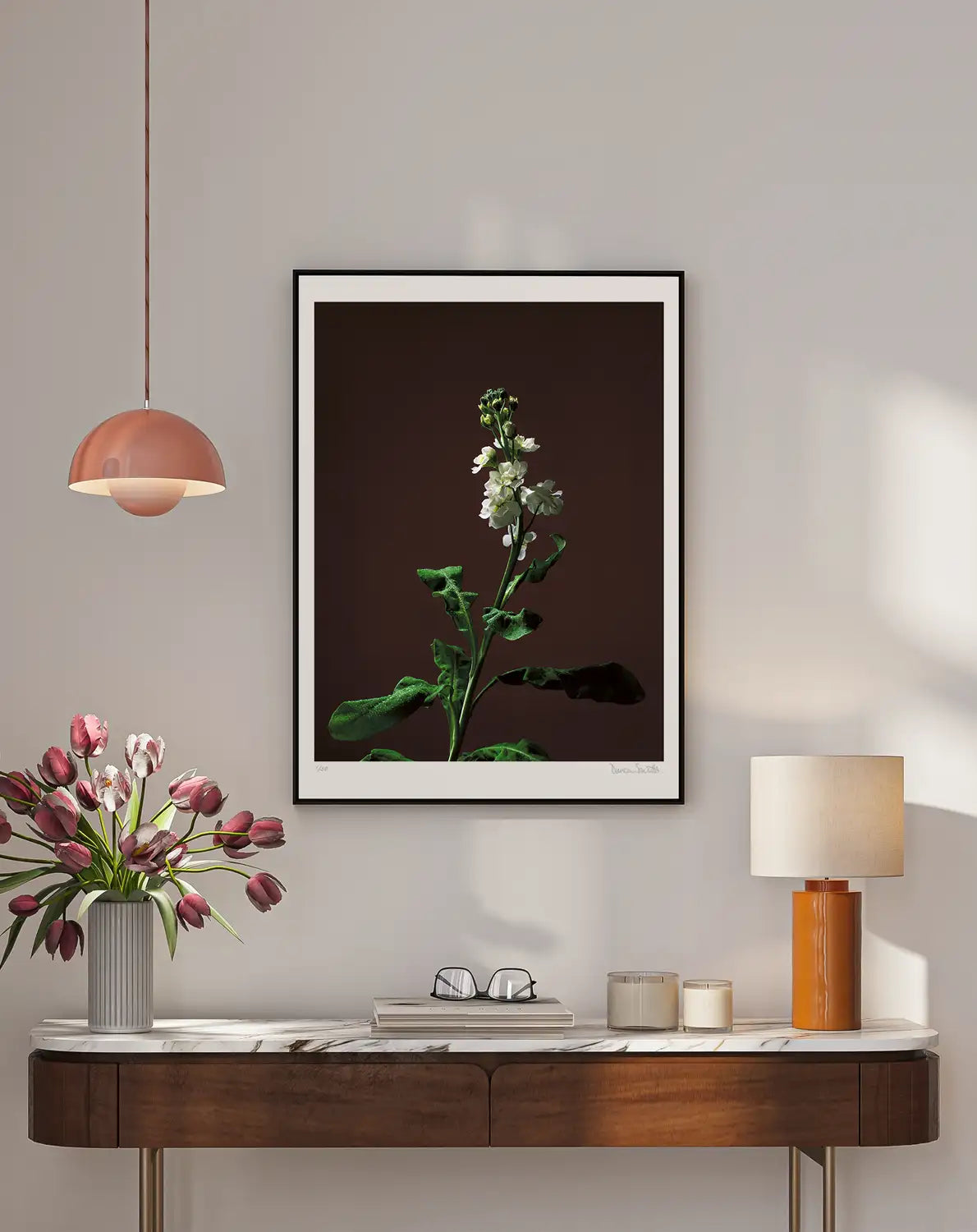 A lifestyle image for representation only featuring a large framed botanical photographic art print featuring a white stock flower on a rich rusty brown background, displayed on a light grey wall above a modern rounded corner wooden console table with a vase of pink tulips, a pinkish hanging pendant light, and a orangish barrel ceramic table lamp. A couple of candles in a jar. A book with glasses finishes the picture.