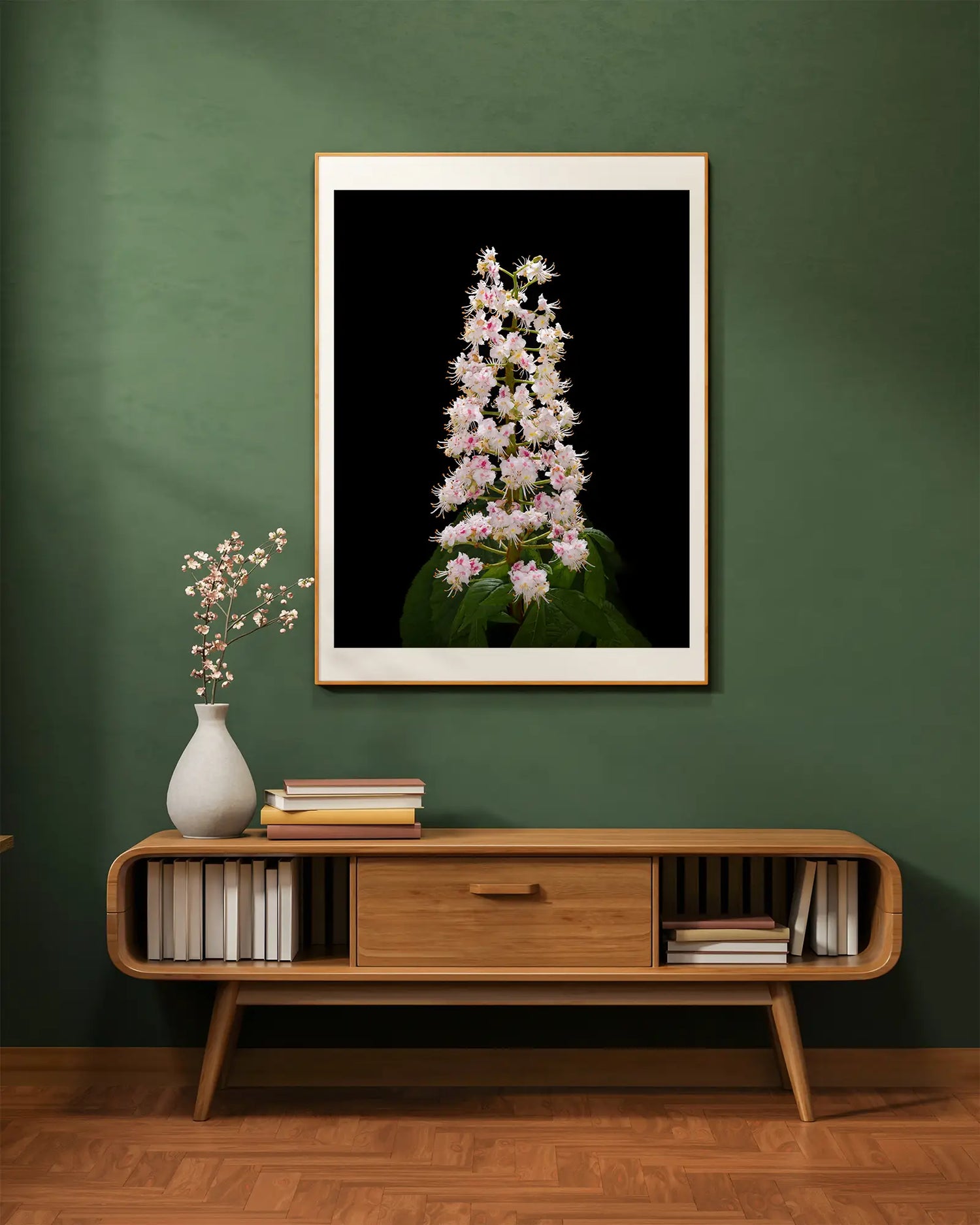 A large framed picture of a horse chestnut blossom 'candle' hanging on a green wall with a bookcase and a vase of flowers below